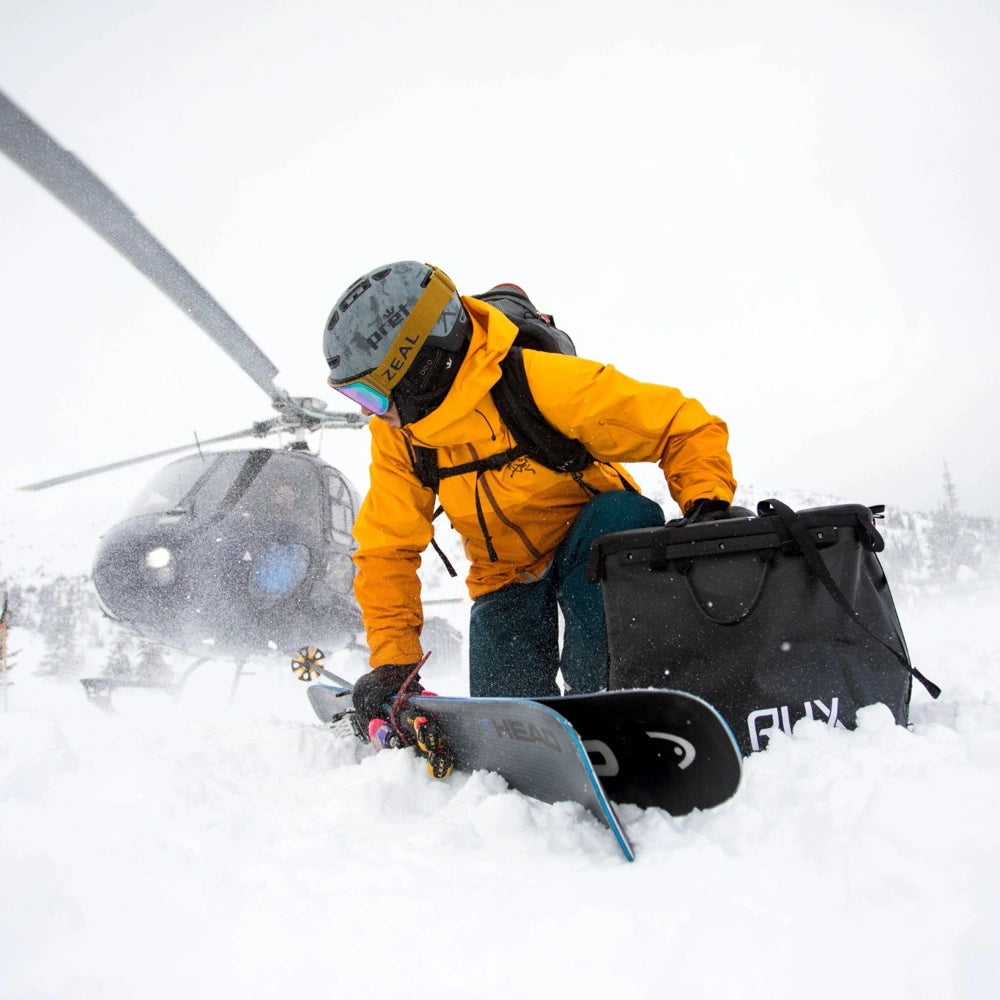 RUX 70L Pack durable gear storage system in snowy backcountry with snowboard and helicopter for skiing adventure and winter outdoor travel