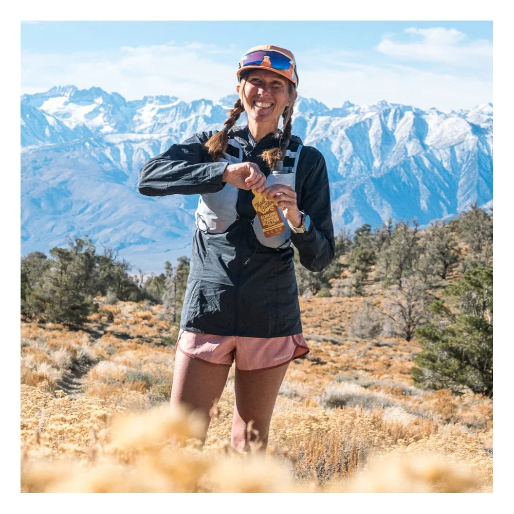 Trail Butter Maple Syrup & Sea Salt Almond Cashew Butter Blend standalone Nutrition Facts panel showing 200 calories per serving, 17g total fat, 5g protein, 8g total carbs, and 160mg sodium per 33g packet
Smiling female trail runner wearing orange visor and running vest squeezing a Trail Butter Almond Cashew Butter Blend Maple Syrup and Sea Salt packet outdoors in a desert scrubland landscape with snow-capped mountain range in the background