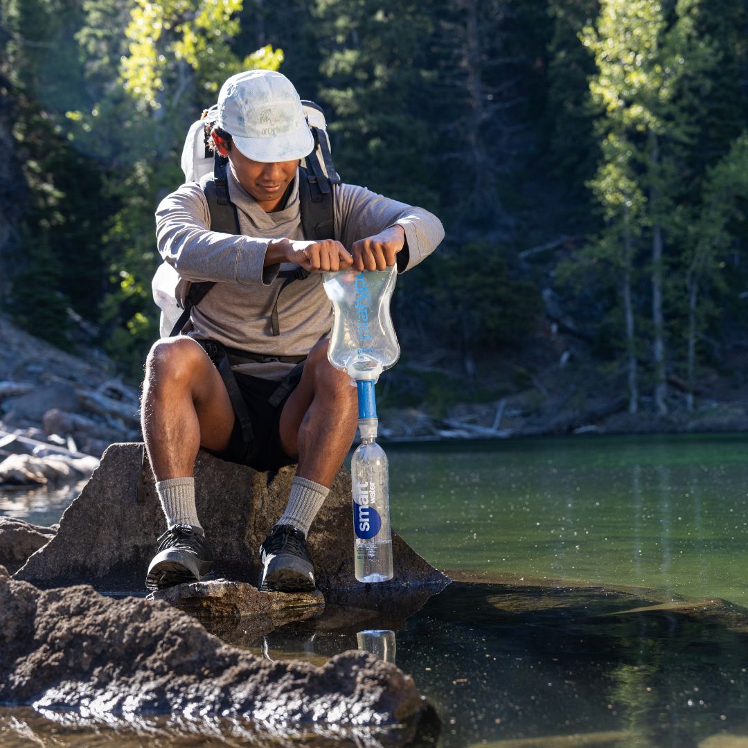 Hiker using platypus quickdraw filter gravity setup filtering water into bottle by lake.