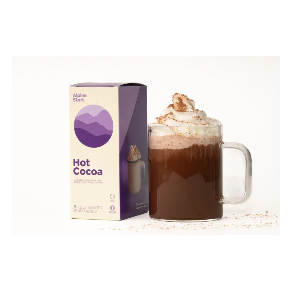 Alpine Start Hot Cocoa 5-packet box next to tall glass mug filled with rich hot cocoa topped with whipped cream and cocoa powder dusting on white background