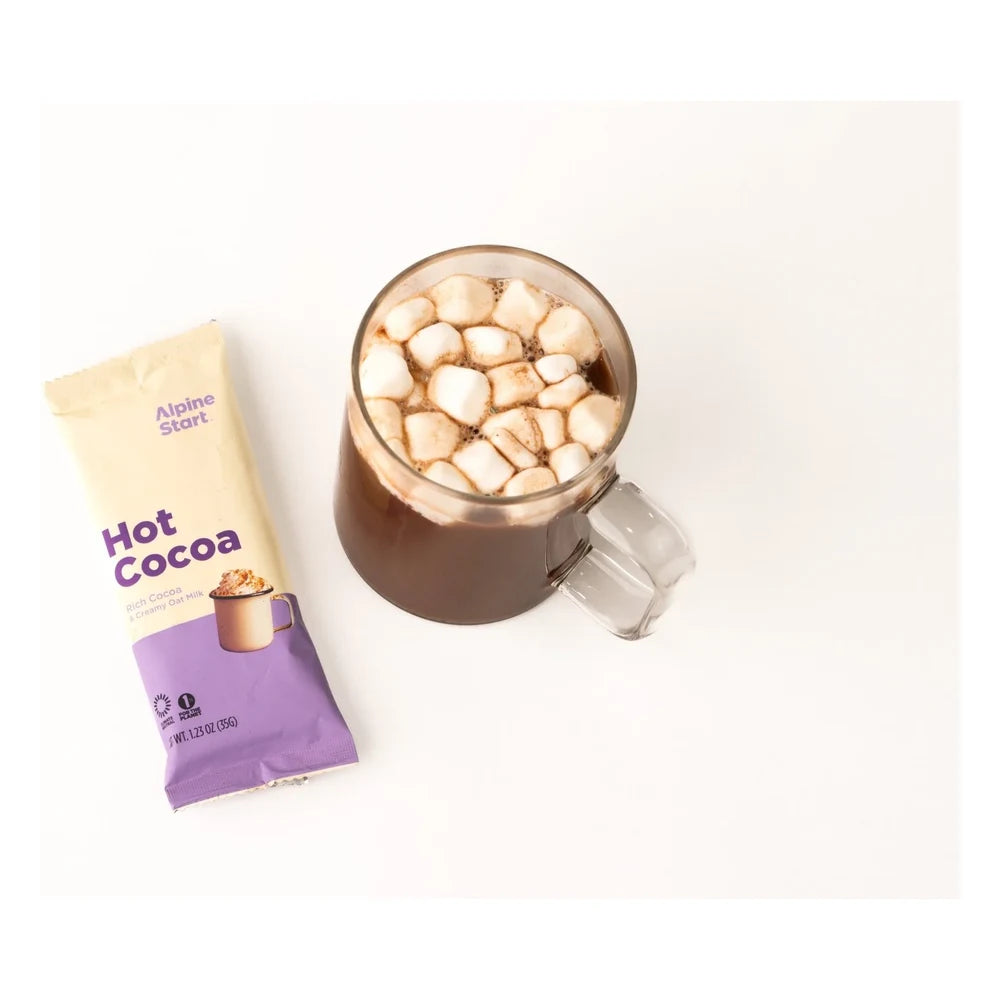 Alpine Start Hot Cocoa single cream and purple stick packet next to glass mug filled with hot cocoa topped with mini marshmallows on white background, showing serving suggestion