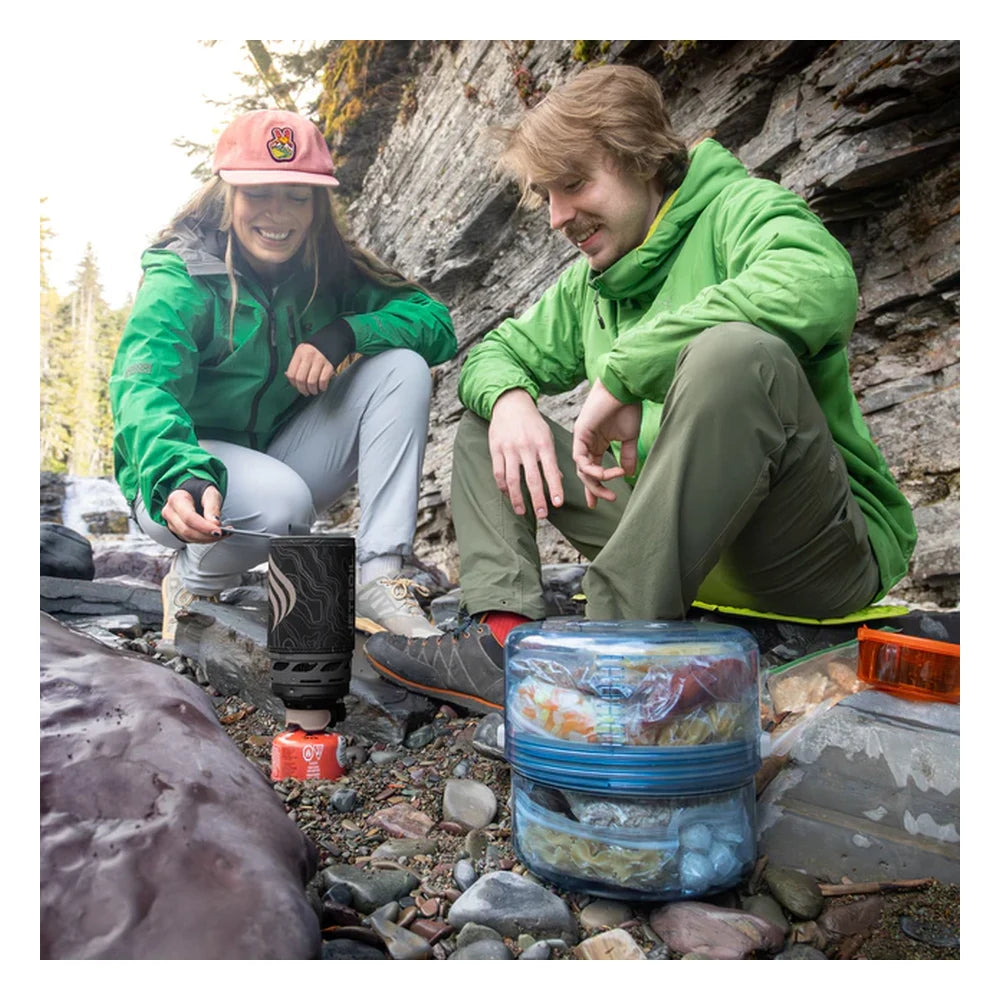 Man and woman cooking at rocky alpine campsite with BVONE collapsible bear canisters in the foreground filled with food, showing real-world backpacking camp use in mountain terrain