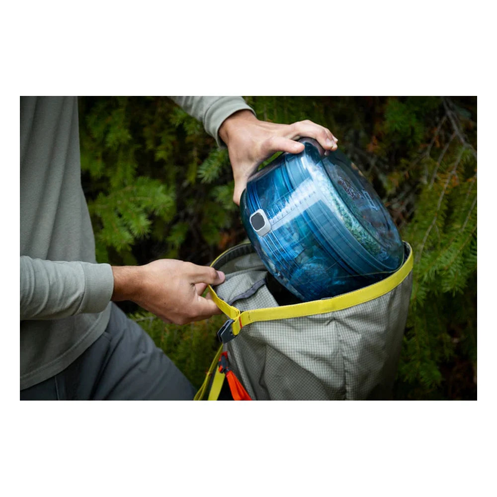 Hiker placing collapsed BVONE bear canister into a backpack in a forest setting, demonstrating compact packable size when collapsed for backpacking and wilderness camping