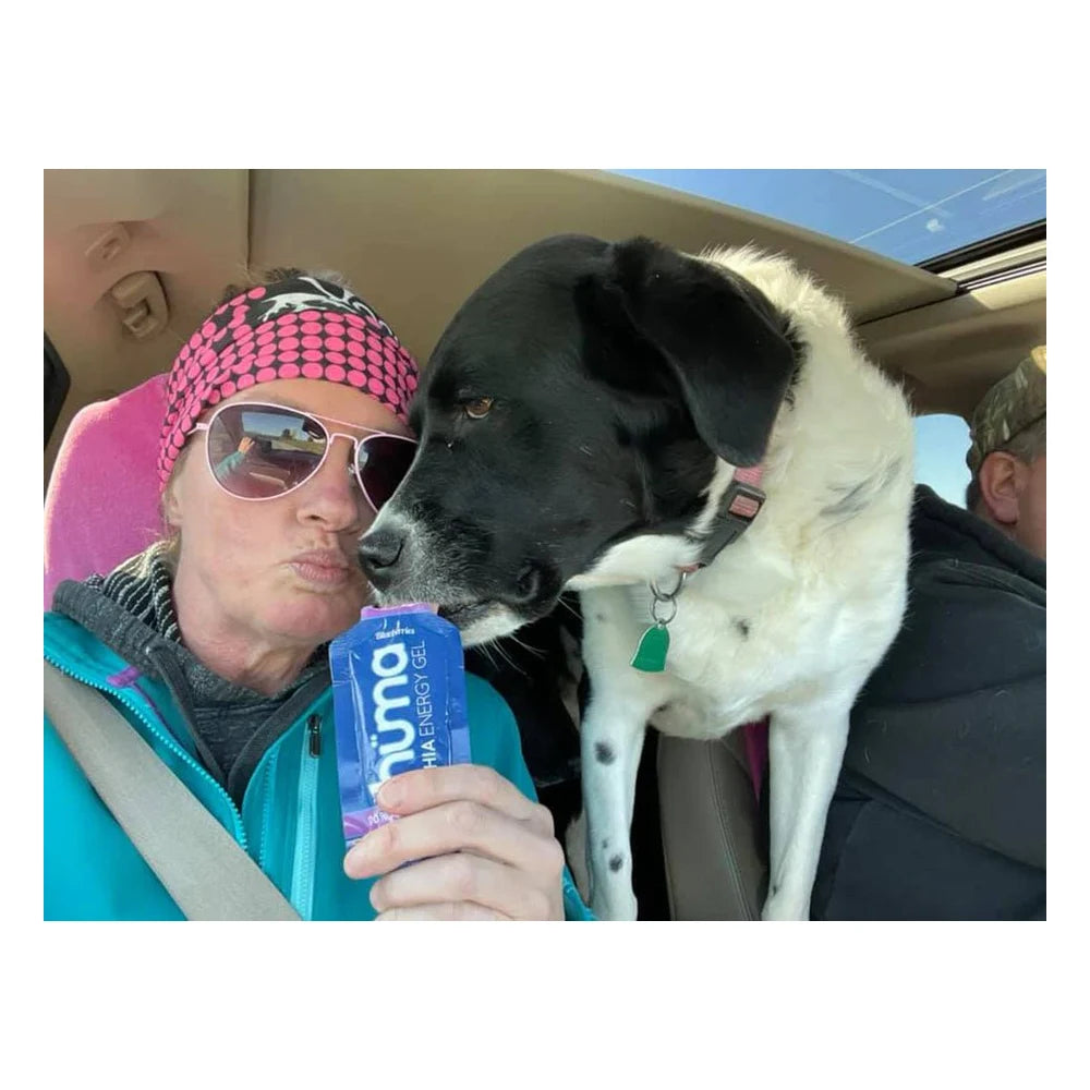 Female athlete in a teal jacket and pink polka-dot headband holding a blue Huma Chia Energy Gel Blueberries packet inside a car while a large black and white dog leans in curiously to sniff it