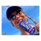 Low-angle selfie of a smiling female runner in a black visor and orange sunglasses biting into a blue Huma Chia Energy Gel Blueberries packet with a bright blue sky in the background