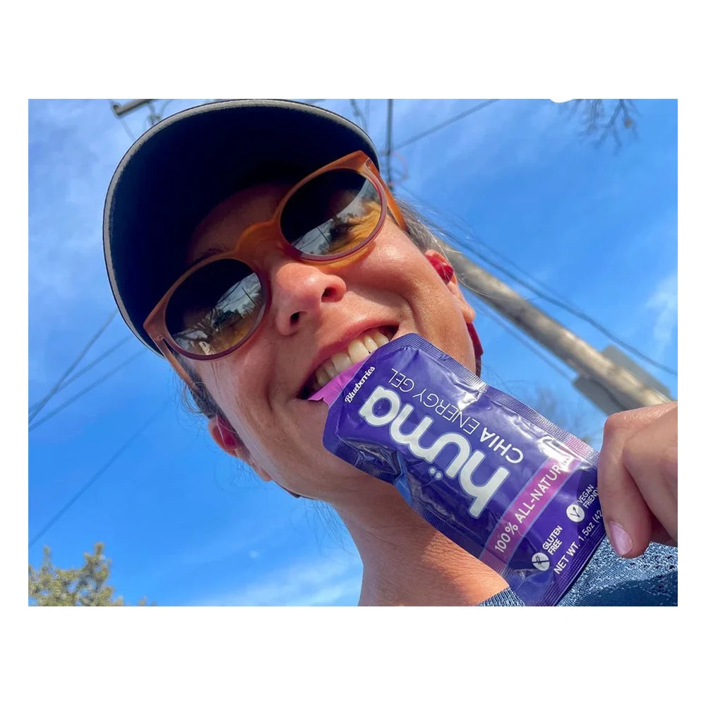 Low-angle selfie of a smiling female runner in a black visor and orange sunglasses biting into a blue Huma Chia Energy Gel Blueberries packet with a bright blue sky in the background