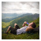 Male hiker lying on his back on a lush grassy alpine ridgeline eating a blue Huma Chia Energy Gel Blueberries packet, with a sweeping panoramic view of green mountain valleys and snow-capped peaks stretching into the distance behind him