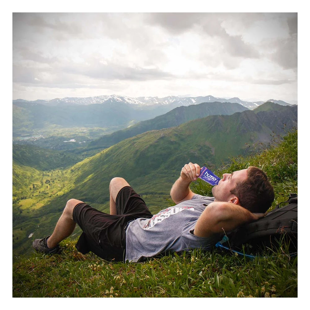 Male hiker lying on his back on a lush grassy alpine ridgeline eating a blue Huma Chia Energy Gel Blueberries packet, with a sweeping panoramic view of green mountain valleys and snow-capped peaks stretching into the distance behind him