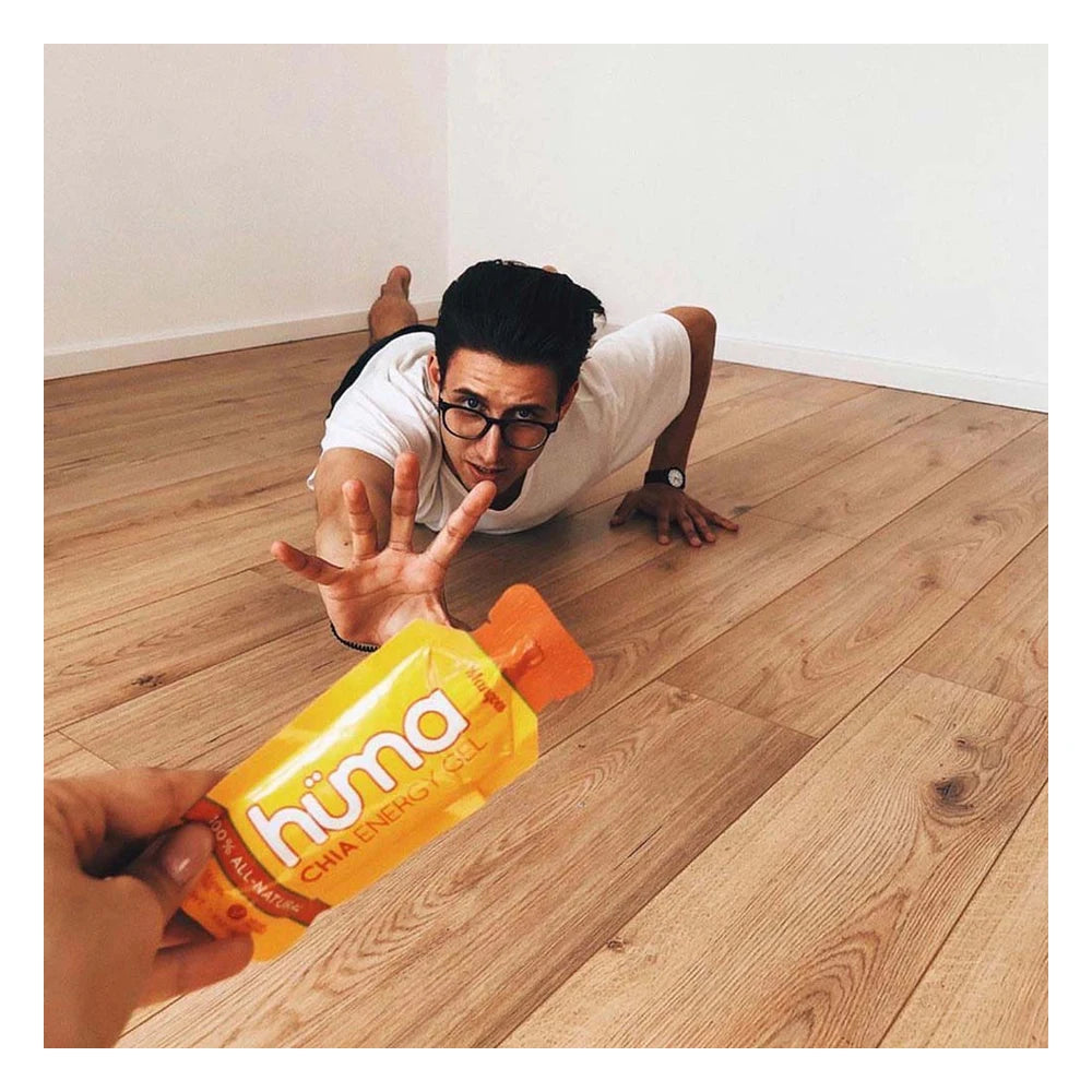 Humorous lifestyle photo of a man in a white t-shirt and glasses doing a low crawl on a hardwood floor reaching eagerly toward a yellow Huma Chia Energy Gel Mangoes packet being held out in the foreground
