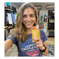 Smiling female runner wearing a Great American Running Show t-shirt holding up a yellow Huma Chia Energy Gel Mangoes packet inside a running specialty retail store with athletic apparel visible in the background