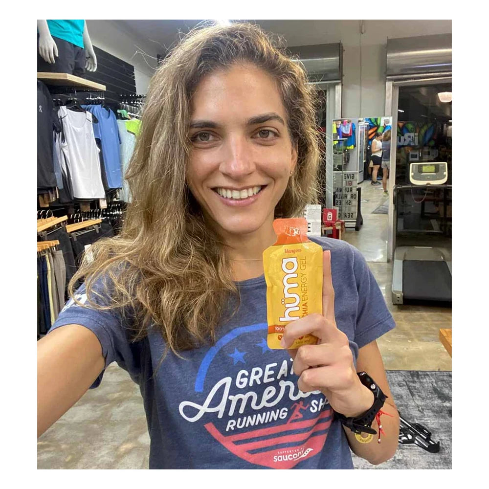 Smiling female runner wearing a Great American Running Show t-shirt holding up a yellow Huma Chia Energy Gel Mangoes packet inside a running specialty retail store with athletic apparel visible in the background