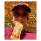Runner selfie outdoors biting open a yellow Huma Chia Energy Gel Mangoes packet wearing a cap and orange sunglasses with vibrant orange and yellow autumn foliage in the background