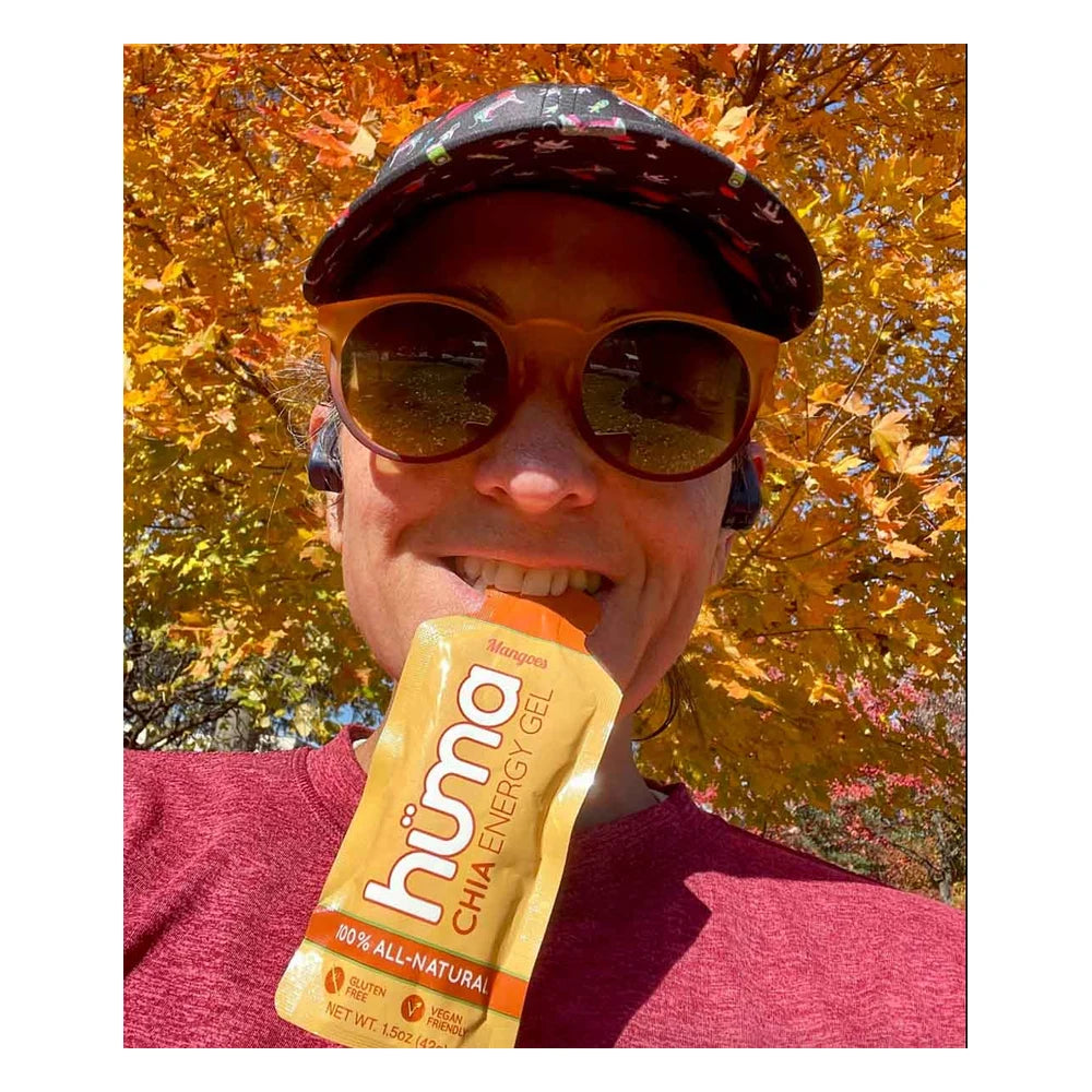Runner selfie outdoors biting open a yellow Huma Chia Energy Gel Mangoes packet wearing a cap and orange sunglasses with vibrant orange and yellow autumn foliage in the background