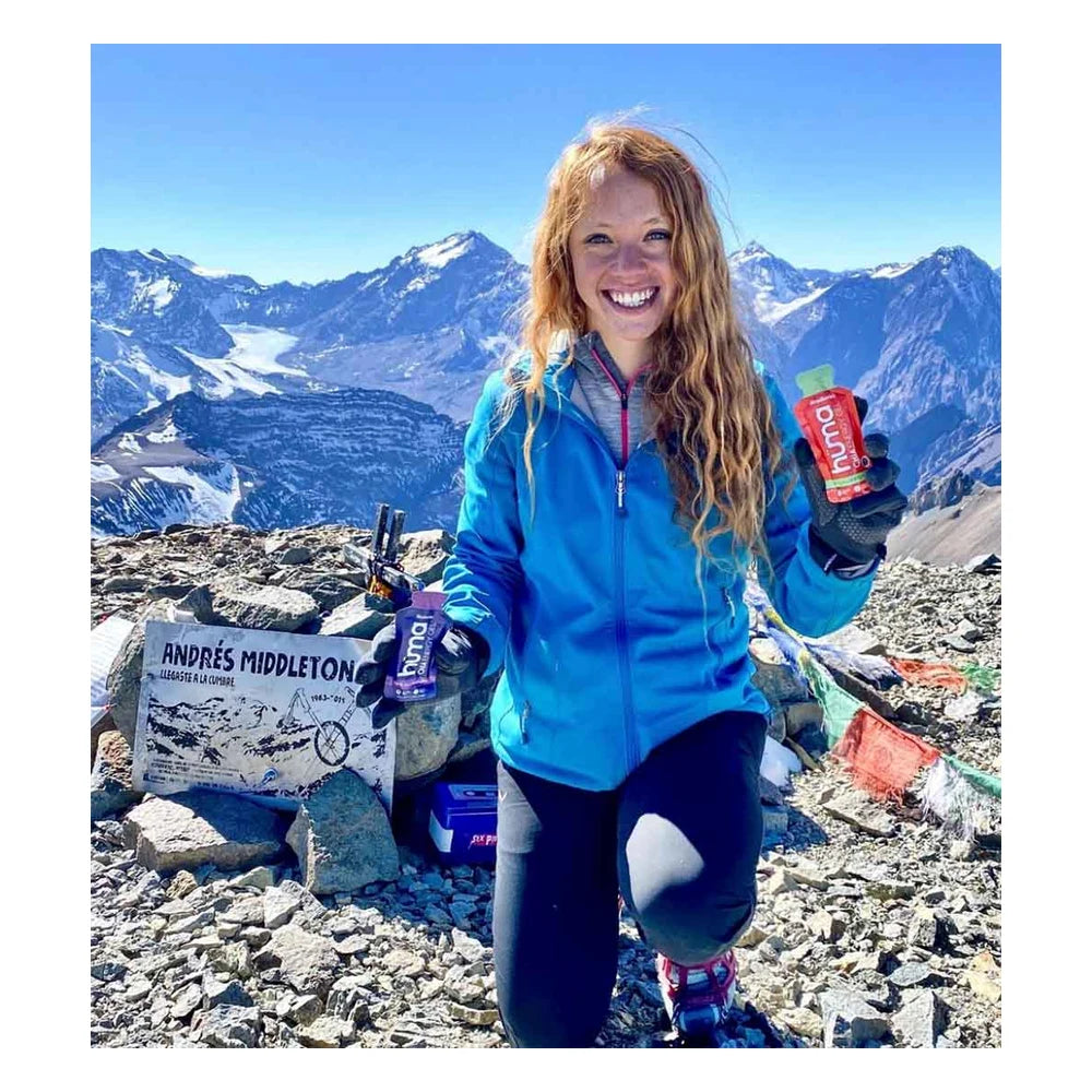 Smiling female athlete in blue jacket holding a red Huma Chia Energy Gel Strawberry packet at a rocky high-altitude mountain summit with sweeping snow-capped peaks and clear blue sky in the background