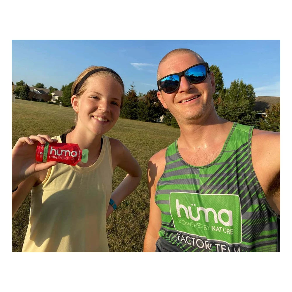 Enthusiastic female athlete wearing a green H√ºma Powered by Nature cap and white sports top laughing with multiple red Huma Chia Energy Gel Strawberry packets taped to her torso and arms, indoor lifestyle setting