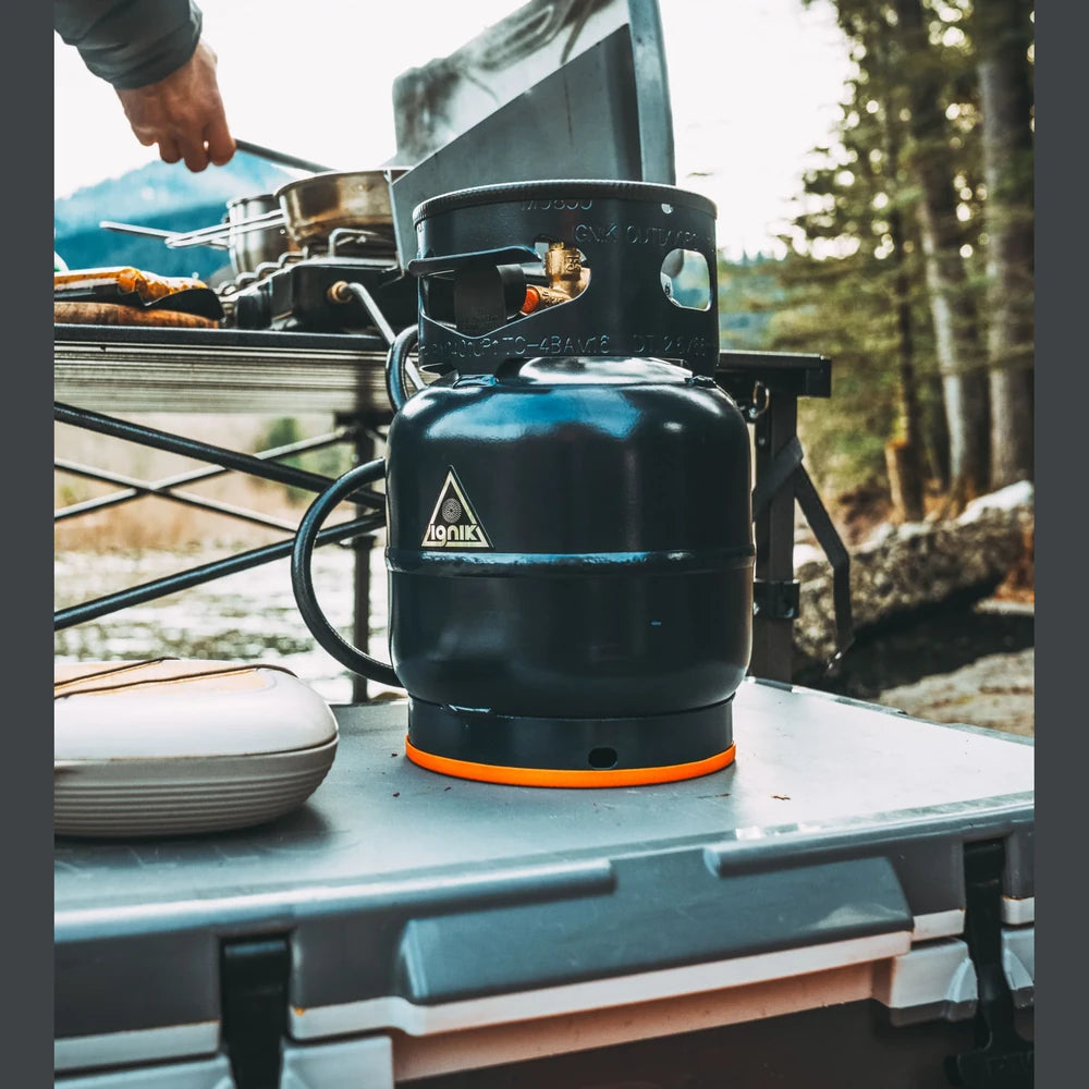 Ignik Propane Growler Tank connected to a camp stove on an outdoor camp kitchen table beside a river