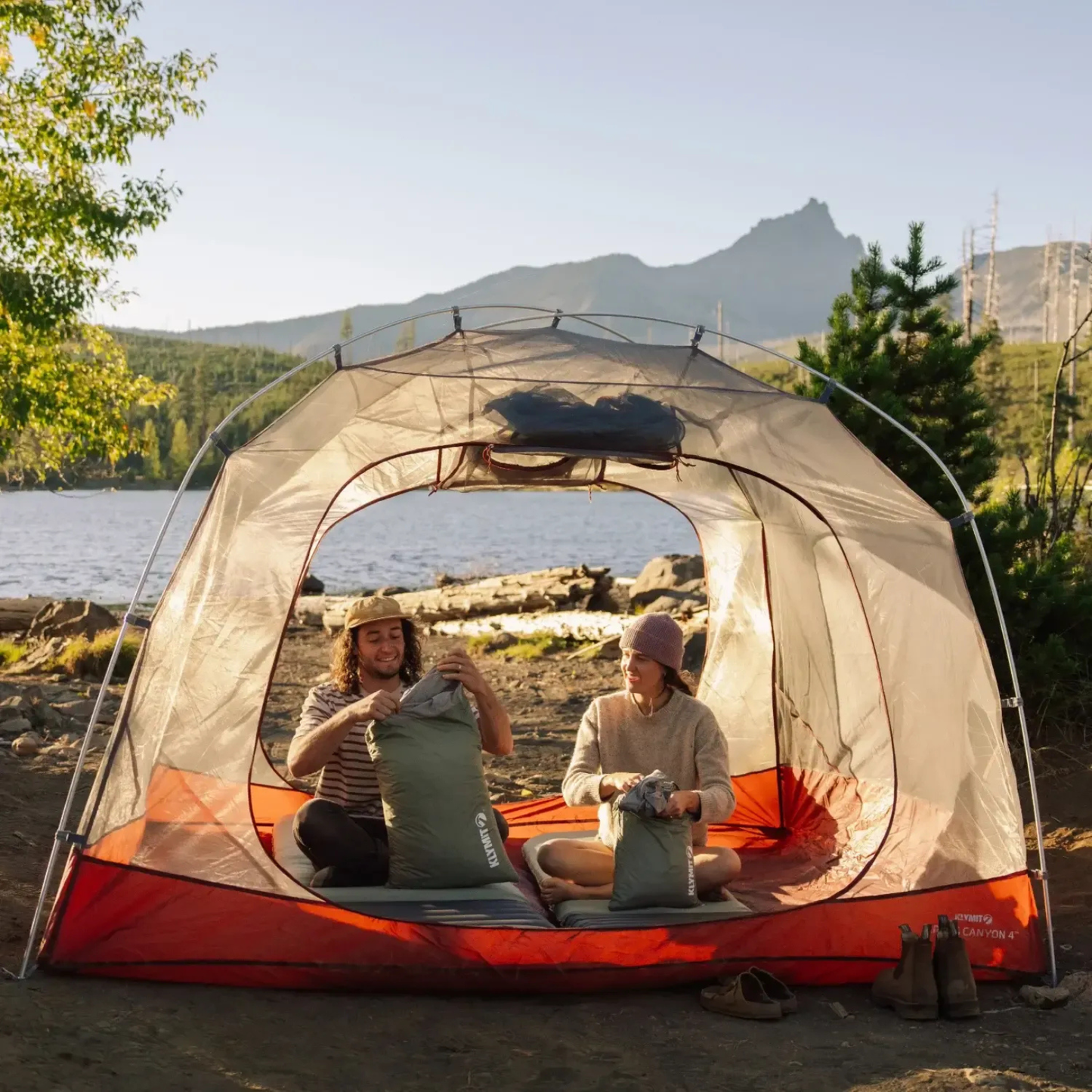 Two campers setting up Klymit sleeping pads inside tent at lakeside campsite in mountains