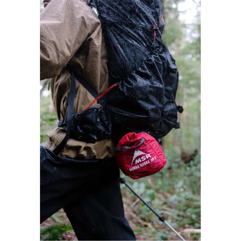Backpacker hiking rainy Vancouver Island trail with MSR Hubba Hubba HD 2-Person Tent stuffsack attached to pack exterior, showing ultralight compact packed size