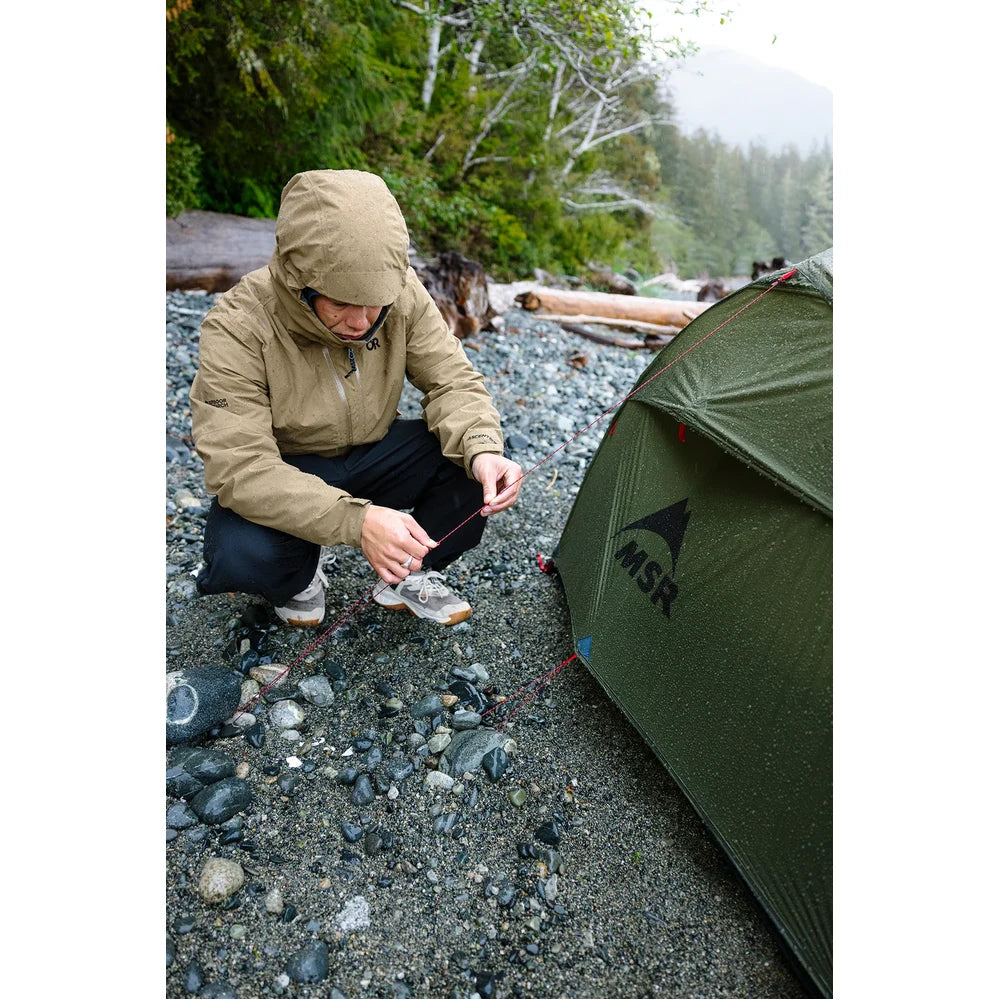 Backpacker staking out MSR Hubba Hubba HD 2-Person Tent on rocky Vancouver Island beach in rainy weather, showing easy pitching and weatherproof olive rainfly