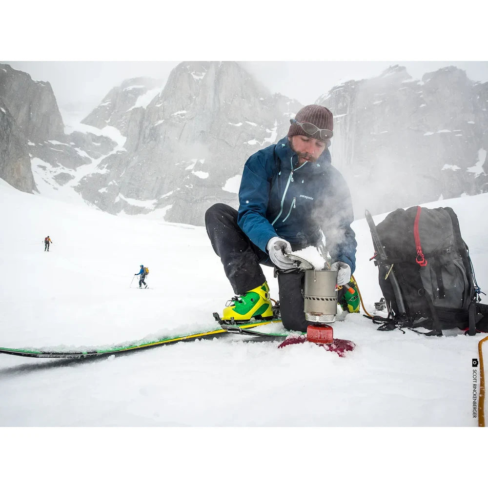 Ski mountaineer melting snow with MSR Reactor stove system in extreme alpine blizzard, demonstrating all-weather radiant burner performance with dramatic spire peaks behind