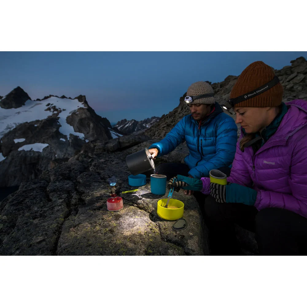 Two backpackers with headlamps cooking with MSR Trail Lite Duo cook set at alpine summit camp at dusk, pouring hot drinks into insulated mugs above snowy mountain peaks