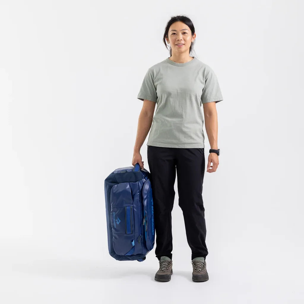 Woman standing and holding a navy Sea to Summit Meridian Duffle Bag by its side handle at her side