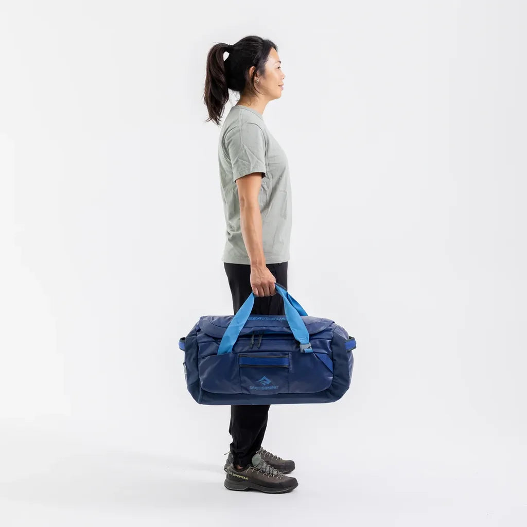 Side profile of a woman carrying the navy Sea to Summit Meridian Duffle Bag by its light blue top handles