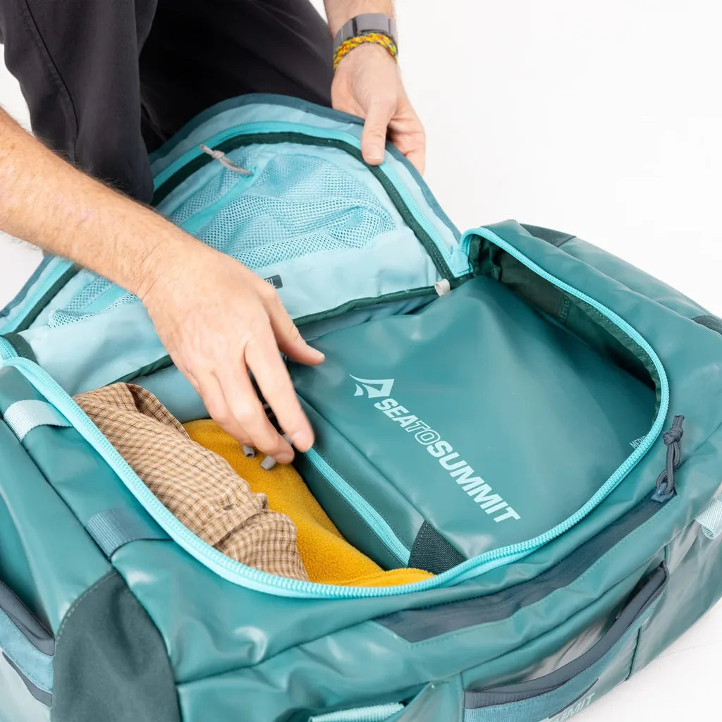 Hands packing clothes into the open main compartment of a teal Sea to Summit Meridian Duffle Bag with mesh pocket in the lid