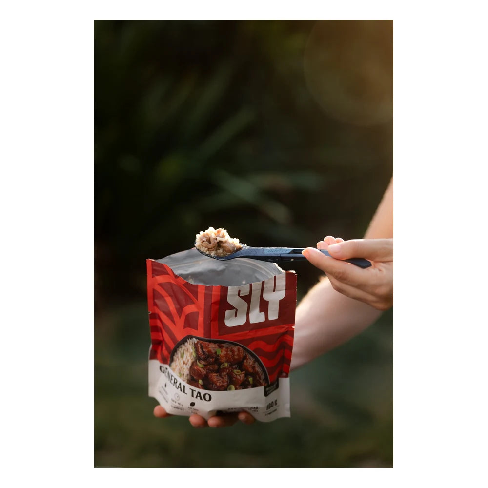 Sly Foods General Tao meal lifestyle shot outdoors showing hands holding the open red SLY General Tao package with a black spork held above the opening, the meal visible inside the open foil bag against a soft-focus green foliage background in warm golden light