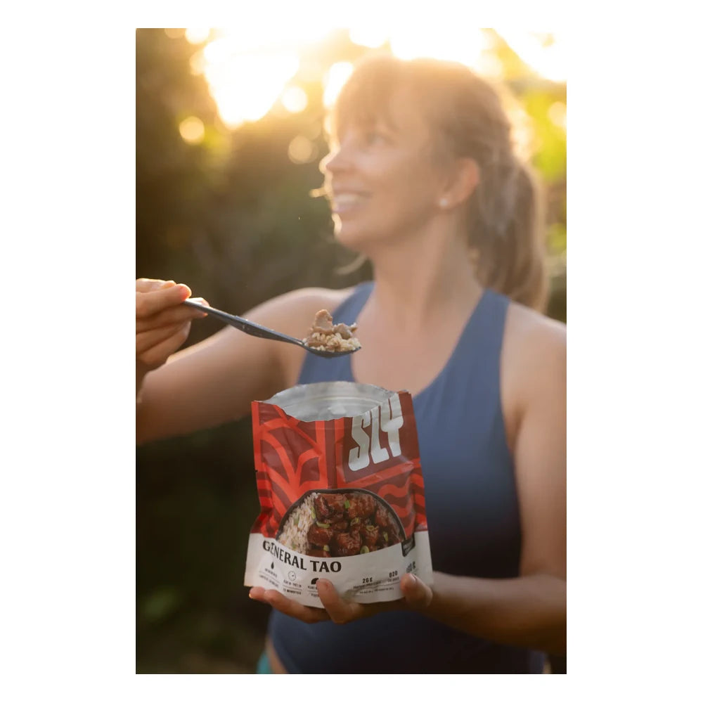 Sly Foods General Tao meal lifestyle shot outdoors showing a smiling woman in a blue tank top eating from a black spork while holding the open red SLY General Tao package, backlit by warm golden hour sunlight with soft green foliage in the background