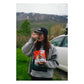 Sly Foods General Tao meal lifestyle shot outdoors showing a smiling woman in a gray sweater and North Face cap eating directly from the open red SLY General Tao package with a spoon, standing in front of a white car with snow-capped mountains in the background