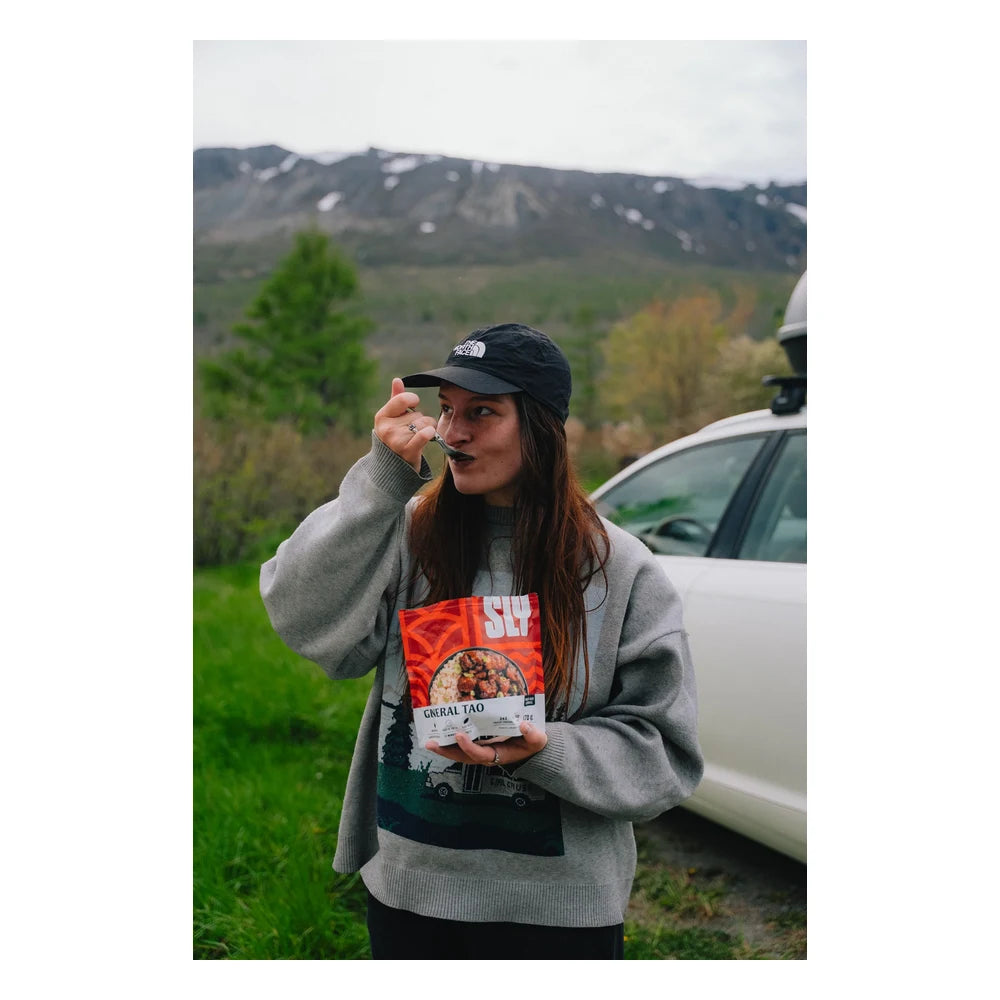 Sly Foods General Tao meal lifestyle shot outdoors showing a smiling woman in a gray sweater and North Face cap eating directly from the open red SLY General Tao package with a spoon, standing in front of a white car with snow-capped mountains in the background