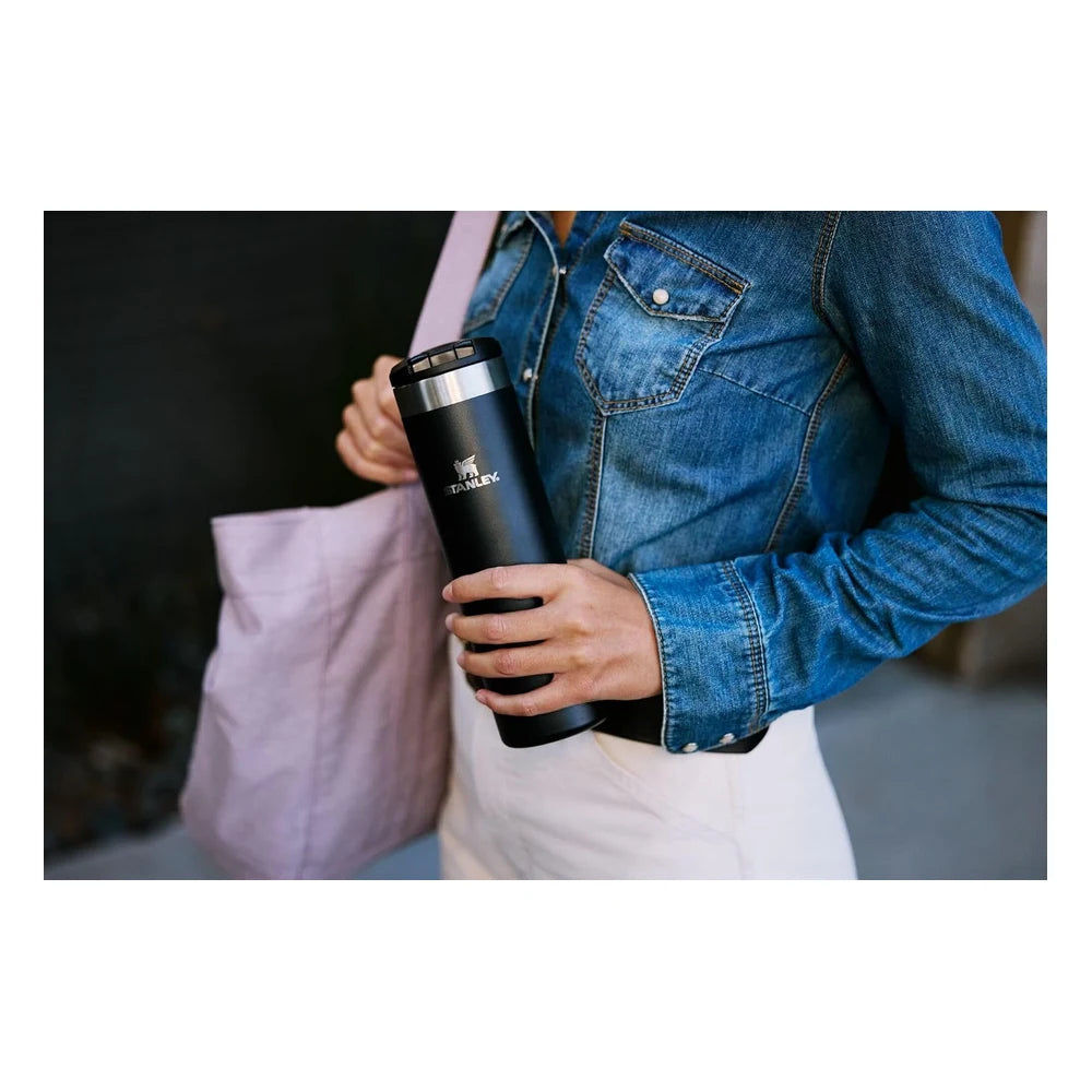 Stanley AeroLight Transit Bottle 20oz in Black lifestyle shot showing a woman in a denim shirt placing the matte black Stanley bottle into a pink tote bag, demonstrating the slim profile fitting easily into a bag