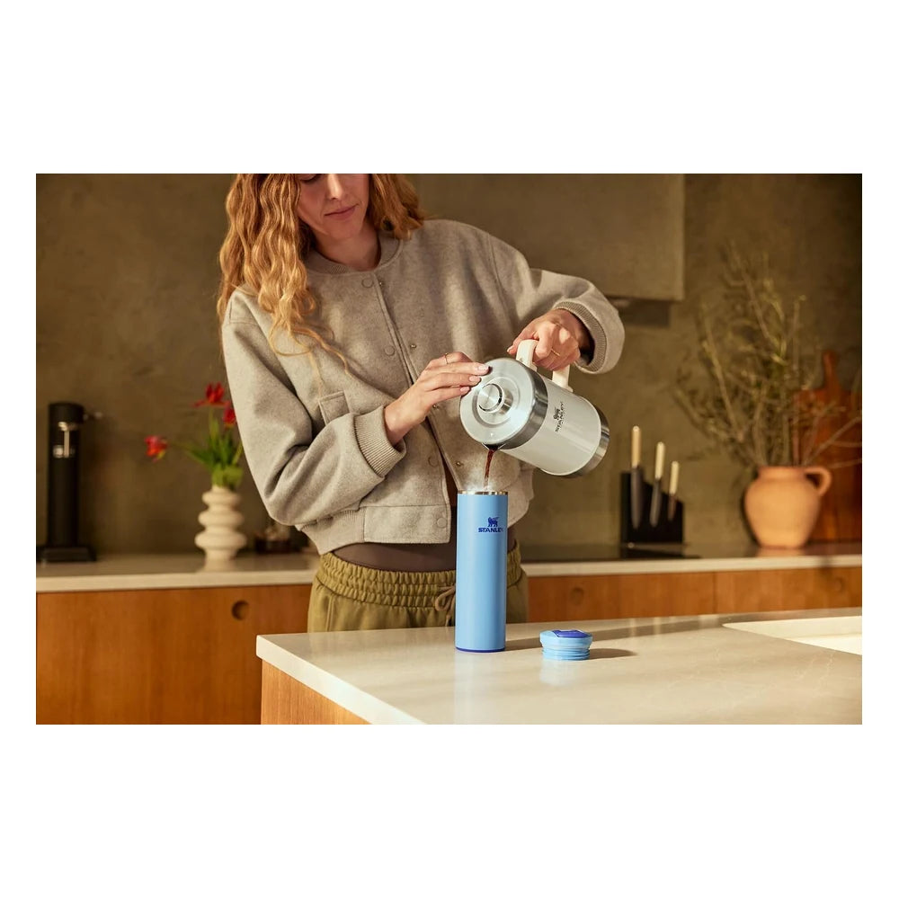 Stanley AeroLight Transit Bottle 20oz in Light Blue lifestyle shot in a modern kitchen showing a woman in a gray cardigan pouring coffee from a white Stanley carafe into the open light blue bottle on a white countertop with the lid set to the side