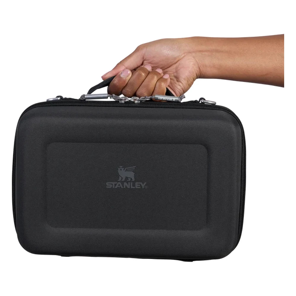 Stanley All Day Midi Lunch Box 6.3QT in Black handle close-up detail on white background showing a hand gripping the molded black top carry handle with silver metal zipper pull hardware, and Stanley winged bear logo embossed on the black textured hard-shell body below