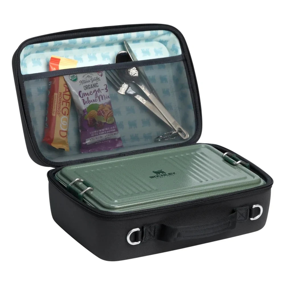 Stanley All Day Midi Lunch Box 6.3QT in Black open view on white background showing the lunch box filled with food and gear: lid organizer pocket holding a snack bar, trail mix bag, and metal utensil set, with a green Stanley food container fitting snugly in the main compartment below