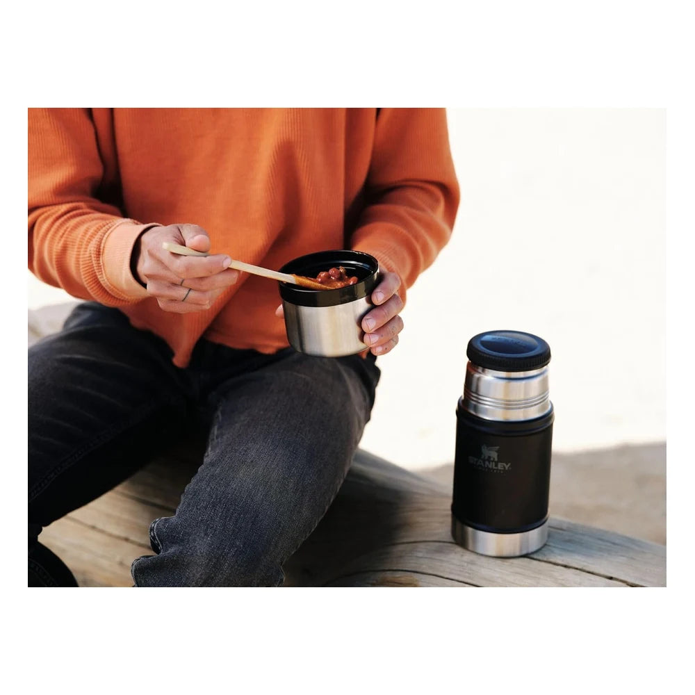 Stanley Classic Legendary Food Jar 24oz in Midnight Black lifestyle shot outdoors showing a person in an orange sweater eating food with a wooden spoon from the open stainless steel inner bowl, with the assembled dark food jar resting on a wooden surface in the background