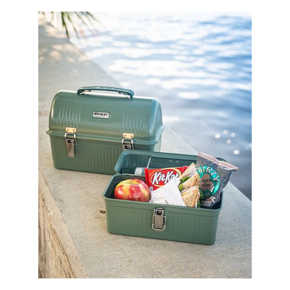 Stanley Classic Lunch Box 5.5QT in Hammertone Green lifestyle shot outdoors on a concrete ledge by the water showing the open lunch box in the foreground filled with an apple, Kit Kat bar, sandwich, and chips bag, with the closed larger lunch box stacked behind under palm tree shade
