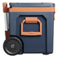 Stanley The Cold For Days Wheeled Outdoor Cooler 50QT in Twilight side view on white background showing the large orange side carry handle with Stanley bear icon and locking release, navy blue body, orange lid trim, and large black all-terrain wheel at the base with the telescoping handle frame visible
