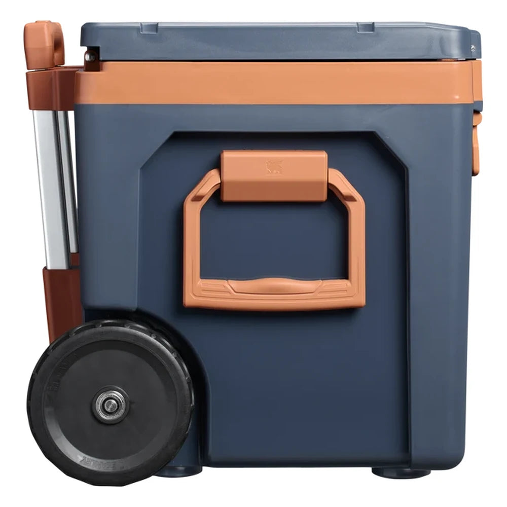Stanley The Cold For Days Wheeled Outdoor Cooler 50QT in Twilight side view on white background showing the large orange side carry handle with Stanley bear icon and locking release, navy blue body, orange lid trim, and large black all-terrain wheel at the base with the telescoping handle frame visible